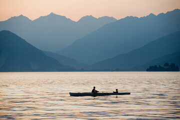 A small rowboat with two people on board sails in the sunset light on the waters of an Italian lake with the silhouettes of the mountains on the horizon