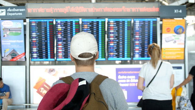 Man in cap with backpack stands back and looks schedule on scoreboard airport. concept of air travel.