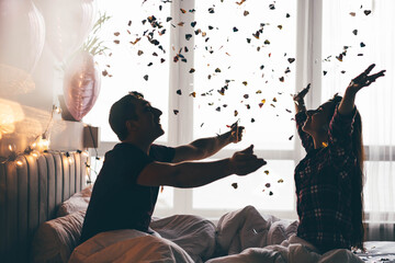 Cheerful young man and woman throw shiny confetti celebrating anniversary in light hotel room...