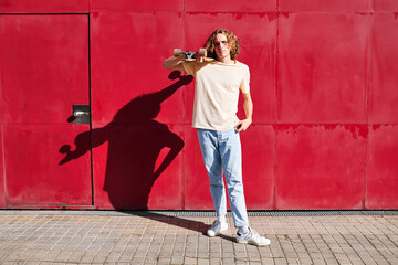 a young man with curly hair and his skateboard with a red background on a sunny summer day