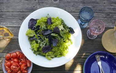 Fresh salad mix of arugula leaves, basil, and lambs lettuce. Salad bowl, healthy food. Bowl of sliced tomatoes and glass of water. Composition in a white plate on an old wooden table
