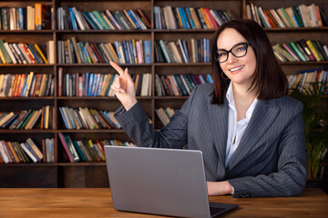 Woman in formal suit sitting at table with laptop pointing to copy space.