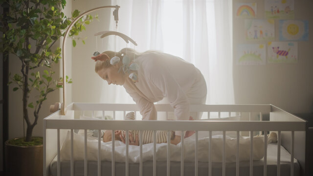 Authentic Shot Of An Adorable Newborn Baby Lying On The Back In Child Crib In Cozy Warm Dark Room. Happy Mother Picks Up Neonate Toddler From The Bed. Concept Of Childhood, New Llife, Parenthood.