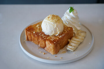 Honey Toast, a square toast baking with butter and topped with vanilla ice cream and honey sauce served with banana and whip cream on white plate.