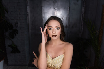 Obraz na płótnie Canvas Close up beauty portrait of girl, holds her hand to head, headache. Brunette with brown eyes, professional evening make-up, dark lipstick, smoky eyes in gold corset. Festive, party. Silver background
