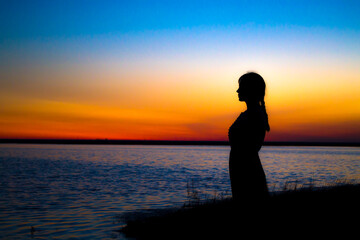 The girl at sunset stands at the shore and looks into the distance, waiting for a miracle.