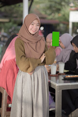 Smiling young Asian teenager girl showing blank green phone screen while friends sitting in background on cafe or eatery