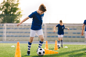 Happy young boy running and kicking the soccer ball in training. Group of school kids practicing football in the outdoor class. Youth soccer player knocking over the training cone.