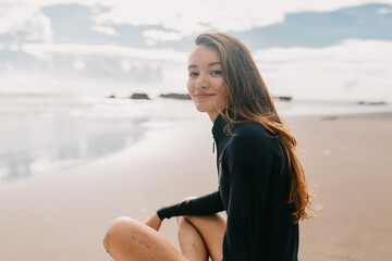 Outdoor portrait of attractive european girl with long hair wearing wetsuit posing at camera near the ocean in warm sunny day