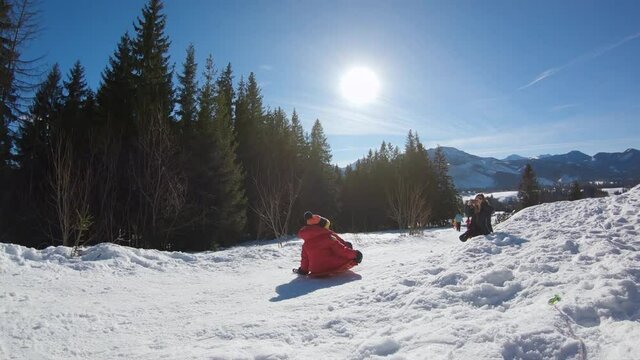 Kid in red jacket jump in slow motion on sled while sledding down from snowy hill in winter during sunny day in Zakopane.
