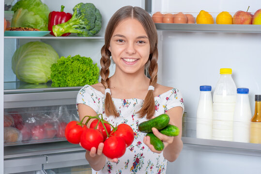 Smiling young teen girl holding fresh red tomatoes and green cucumbers while standing near open fridge in kitchen at home. Portrait of child choosing food in refrigerator full of healthy products
