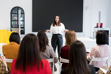 Pretty woman speaker giving talk and gesturing with hands in front of the audience. Coach explaining to group of people at  business meeting. Rear view of  group session in the conference hall