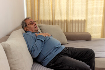 Obraz premium Shot of an uncomfortable looking old man holding his chest in discomfort while being seated on a couch at home. Overweight senior man holding breast because of heart infarction. Health care concept.