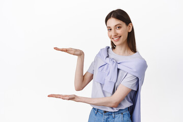 Place your logo here. Smiling cute girl holding empty space between hands, hold box on copy space, showing big size object, standing over white background advertising