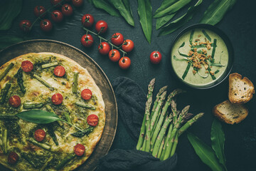 Homemade tarte flambee with green asparagus and tomatoes and wild garlic soup on dark background