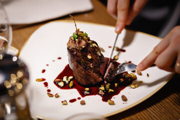 Fork and knife in the hands of a man cuts meat, oval slice of roasted beef sprinkled with pistachios and sprinkled with sauce. Well-done meat nicely served in a white plate restaurant.