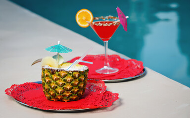 Refreshing cold summer cocktail in a half of pineapple with a straw on a plate on the background of the pool and cocktail sex on the beach. - Powered by Adobe