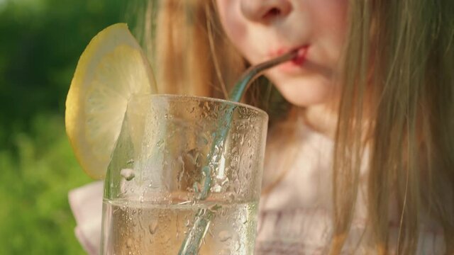 Close-up of unrecognizable child drinking lemonade from glass through reusable metal straw. Little girl with long blond hair quenches her thirst on hot summer day outdoors, selective focus.