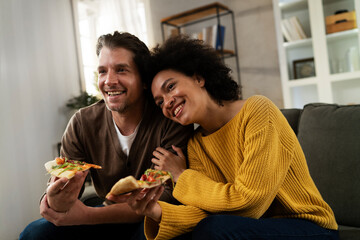 Cheerful young couple sitting on sofa at home. Happy woman and man eating pizza while watching a movie.