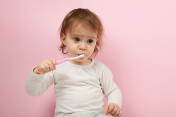 Cute baby girl brushing teeth on pink background.