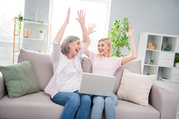 Photo of triumphant old woman and blonde young lady raise hands winner hold computer indoors inside house