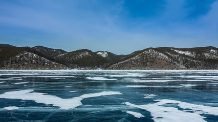 On the smooth ice of the frozen lake, patches of snow and cracks are visible. On the shore, against the background of a blue sky, there is a wooded mountain range covered with snow. Baikal