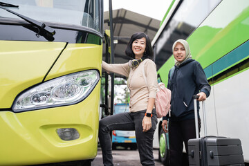 Naklejka premium close up of a short-haired woman with headphones and a bag smiling at the camera as she steps up to the bus door with a woman in a veil carrying a suitcase behind her when going on the bus