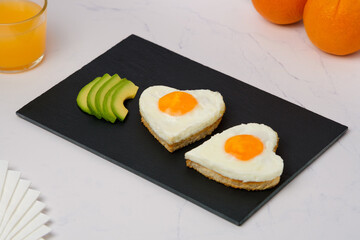 Breakfast.Heart shaped fried egg sandwiches with avocado slices on a stone plate and orange juice.