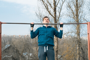 Young man in sportswear exercising outside, pulling himself up on a horizontal bar. Power workout in the fresh air