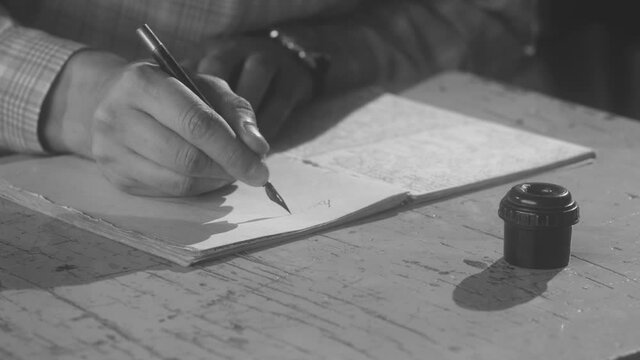 Man siting at a desk and using retro fountain pen and inks to make notes. Stock footage. Close up of male hand dipping his pen into inks and starting to write something in a paper notebook, vintage