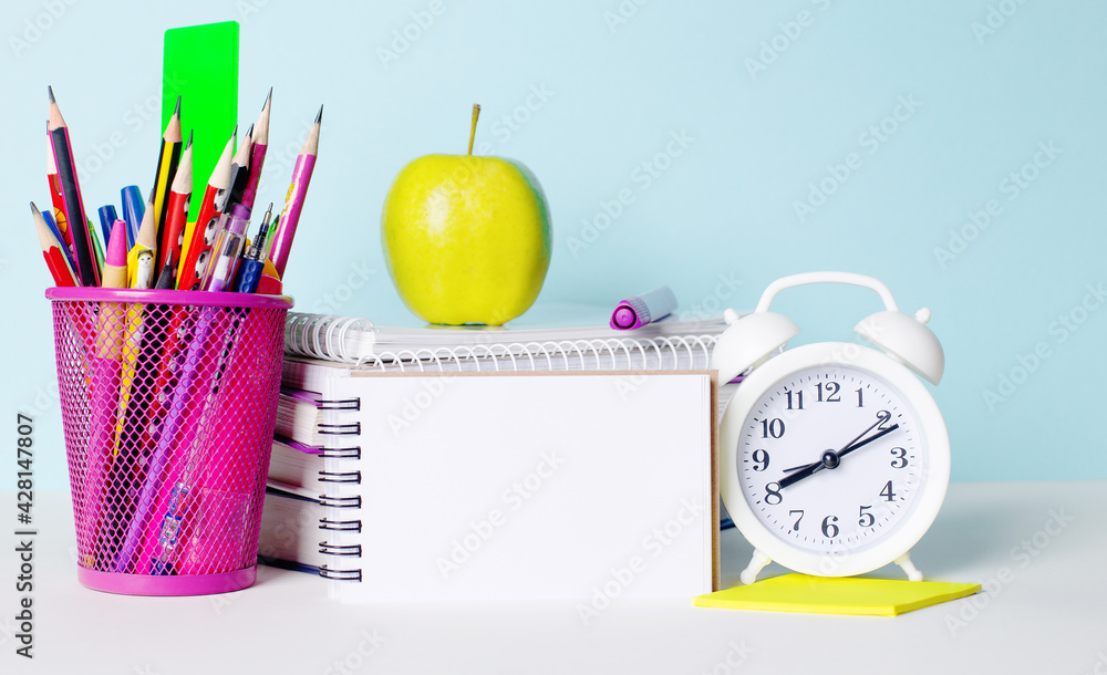 Poster on a light table are books, stationery, a white alarm clock, an apple. next to a blank notebook with