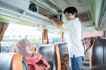 a young man smiles as he helps a woman in a headscarf put her bag on a shelf while standing on the bus