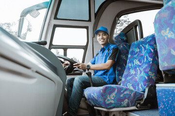 A male driver in a uniform and hat smiles at the camera while sitting on the bus