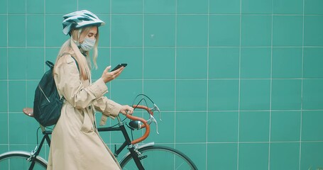 Woman in protective mask in helmet using her smartphone device. Female go to work on his sport retro bike. Girl standing on green background. - Powered by Adobe
