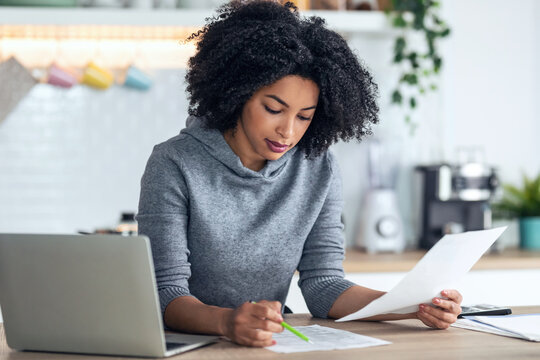 Afro business woman working with computer while consulting some invoices and documents in the kitchen at home.