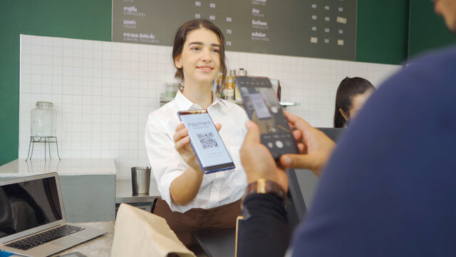Young Middle East woman barista people in apron collecting an order payment by scanning QR code on phone screen, talking to customer at the counter, coffee shop cafe cashier. People lifestyle. Service - Powered by Adobe