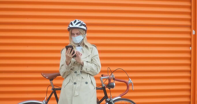 Woman in helmet using her smartphone device. Female his sport bike. Girl standing on orange background.
