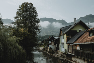 Fototapeta na wymiar Mountain river and row of houses near it in Slovenian town Bohinjska Bistrica under the Julian Alps hiding in clouds in the background during the sunrise during the summer morning