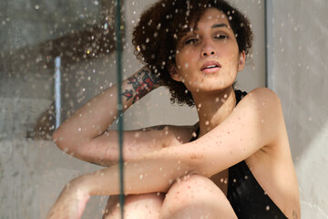 Obraz premium Sensual portrait of young woman taking a shower. Defocused female looks through the glass of the shower stall. Art portrait. Feminine beauty. Skincare wellness.