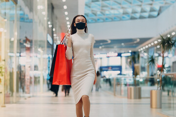 Young adult woman in protective medical face mask carrying paper shopping bags in hands at retail store