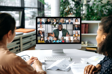 Virtual communication, video call. View over the shoulders of two women to a computer screen with successful multiracial business team gathered in a video conference to discuss working issues,strategy