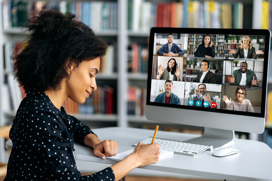 Video call, online conference using app and computer. African American female student, studying remotely, watches an online lecture, taking notes, multiracial smiling people on the computer screen