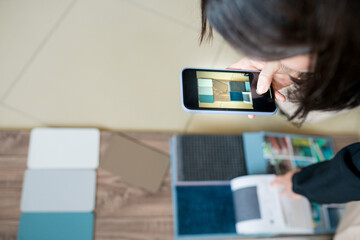 Interior designer using mobile while choosing materials for home renovation. Woman taking photo of samples on the floor and matching the color of the walls to the parquet. House decoration concept