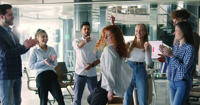 Cheerful young woman startup team leader doing funny dance during break in modern office. Company coworkers having fun at work, celebrating success, expressing satisfection.