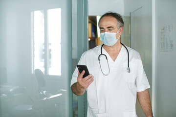 Medico cinquantenne in camice bianco e mascherina chirurgica guarda il suo cellulare  nella corsia...