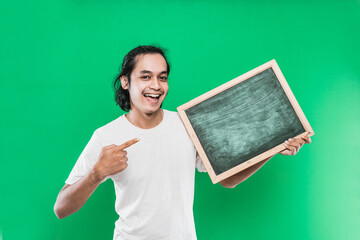 Young man long black hair holding and pointing at something on a blank blackboard with smiling happiness on a green background