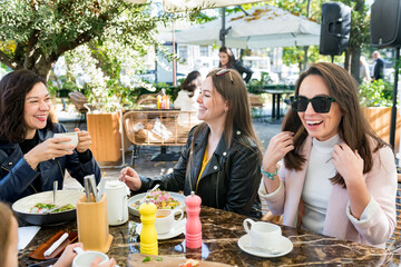 Young happy brunette women friends in casual clothes having fun and laughing during brunch in street cafe on sunny day. Outdoor dining. Selective focus