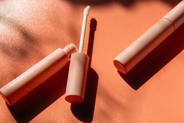 open tube with a brush for liquid lip gloss, liquid lipstick next to closed pink tubes with mascara and eyeliner on a peach background with shadows