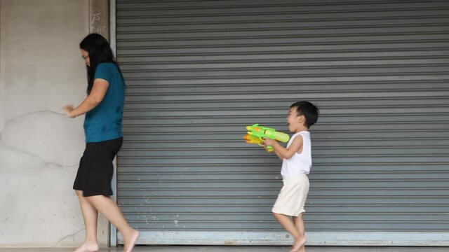 4K Asian child boy playing water gun with mother at home in Songkran day. Concept of happy family activity at home, freedom fun, togetherness. Songkran festival day 2021, new normal Covid 19.
