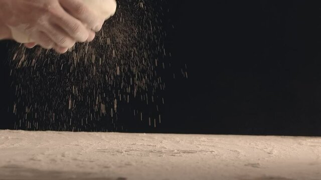 Male hands throw a piece of dough to a wooden table and sprinkle it with flour. Filmed on black background